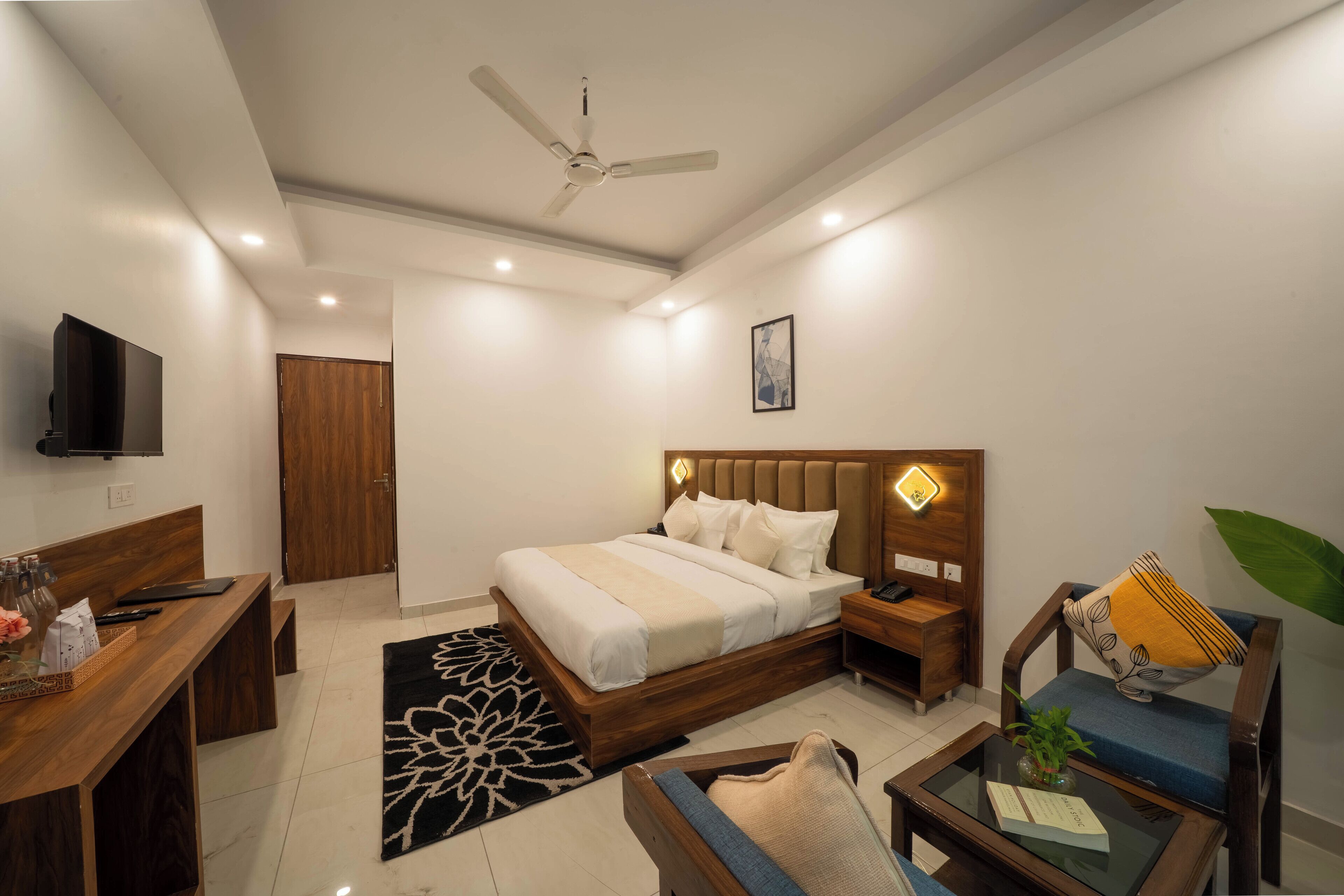 A modern hotel room with a bed, bedside tables, a desk, a chair, a TV, and a ceiling fan.
