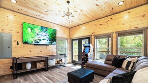 Een gezellige woonkamer met een grote flatscreen-tv, een hoekbank, een poef en een salontafel. Er is een houten plafond, een plafondlamp en een raam met uitzicht op bomen.
