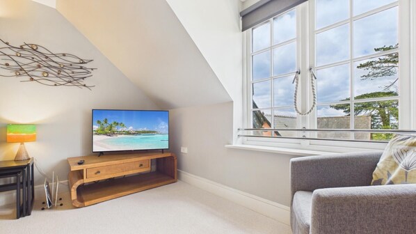 Una habitación con una televisión sobre un soporte de madera, una ventana que da a vistas del cielo y árboles, y un sofá gris.