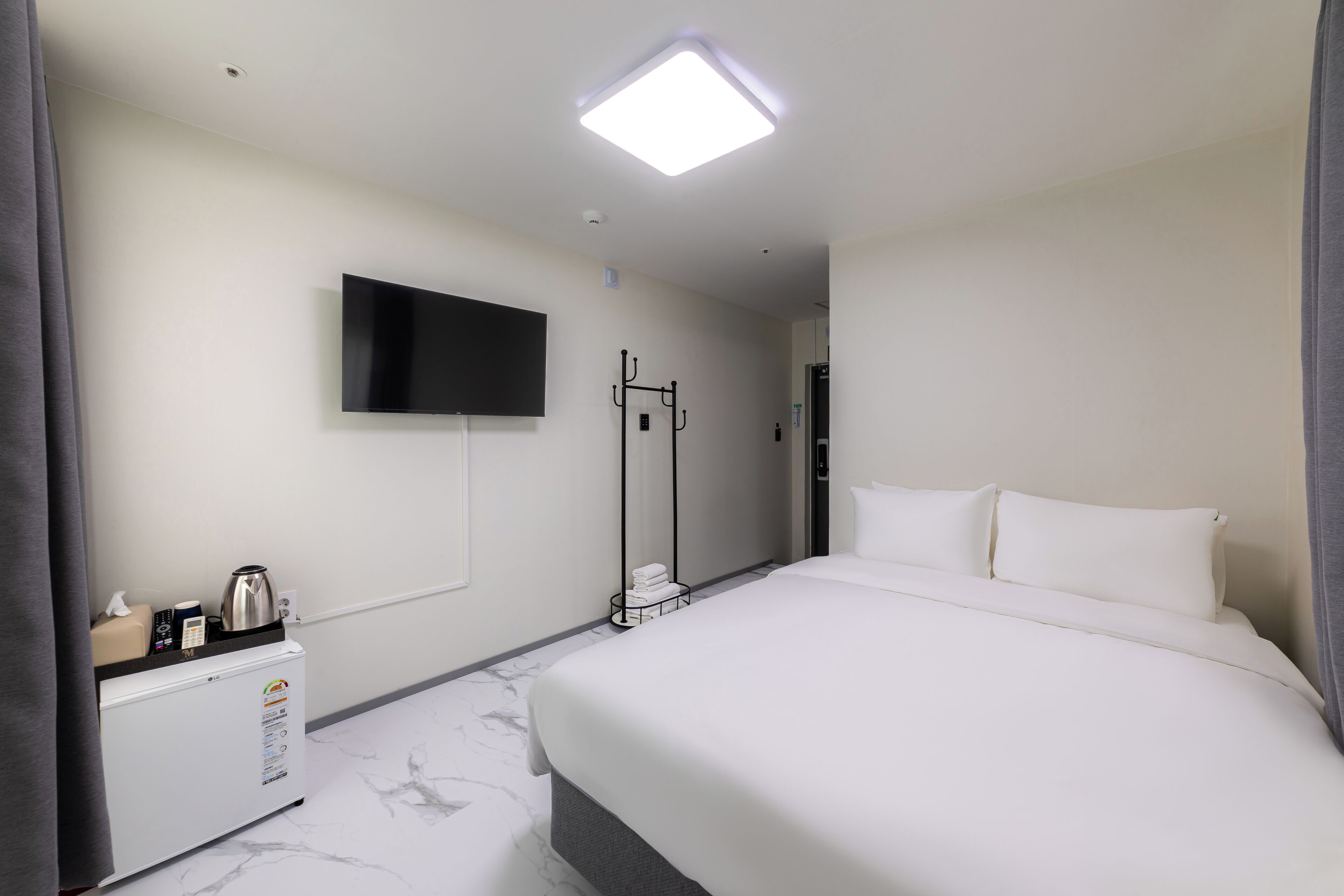 A hotel room with a bed, a TV mounted on the wall, a coffee machine, and a small table with a kettle and a tissue box.