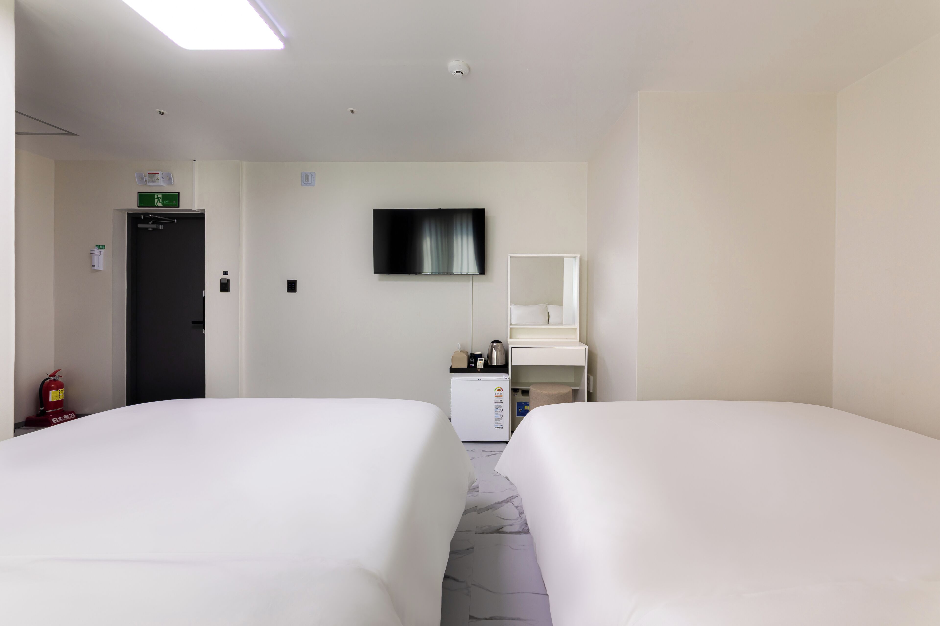 A hotel room with two beds, a television, a small desk with a mirror, a refrigerator, and a fire extinguisher.