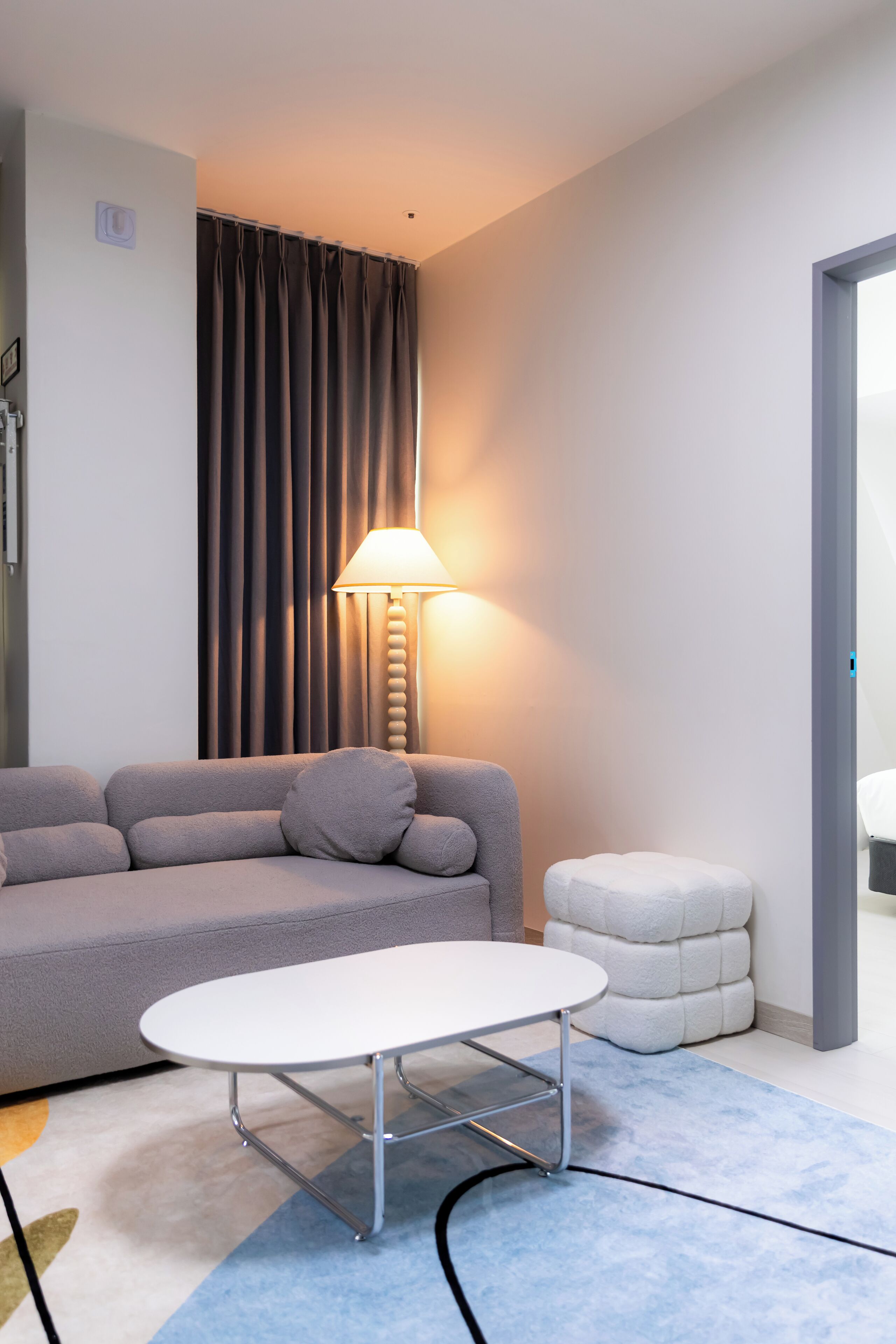 Une chambre d’hôtel moderne avec un canapé gris, une table blanche, une lampe et un petit pouf.
