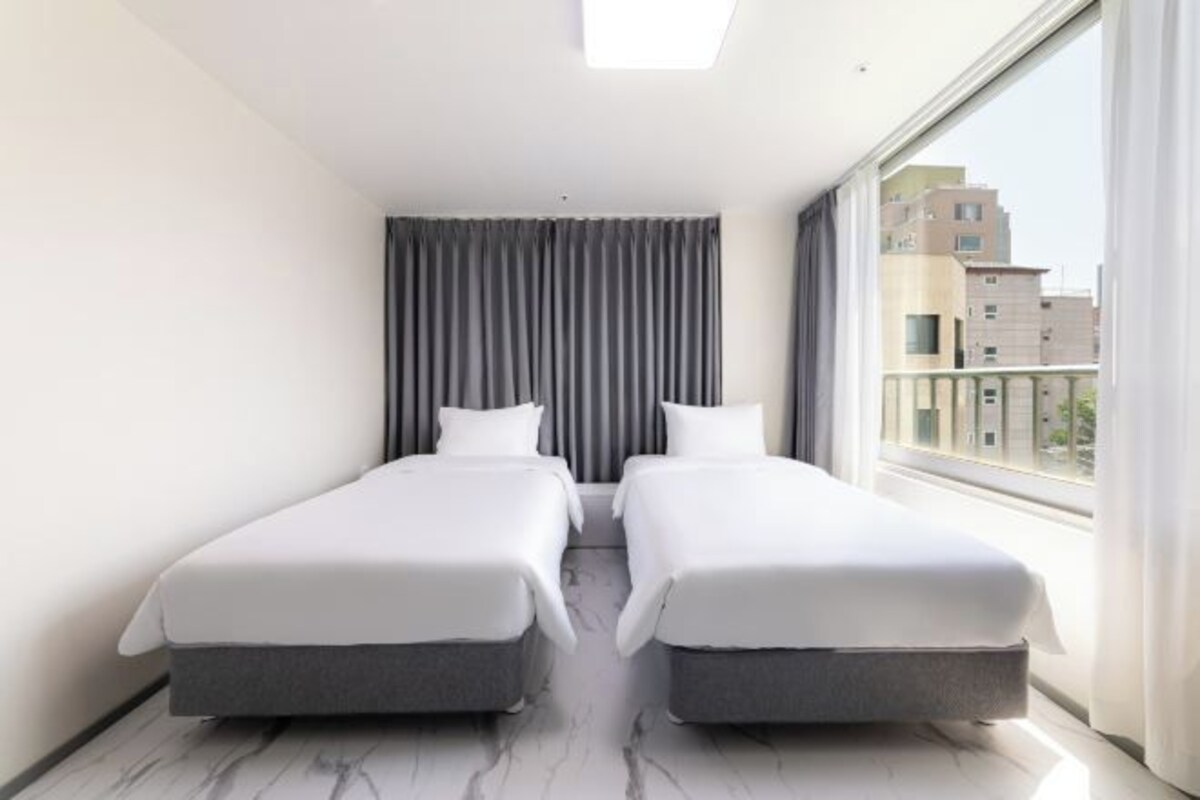 A modern hotel room with two single beds, a large window with curtains, and a view of buildings outside.