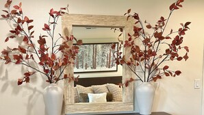 Une commode en bois foncé, sur laquelle reposent deux vases blancs contenant des feuilles rouges. Derrière la commode, on aperçoit un miroir qui reflète un lit recouvert d’une couette blanche et marron.
