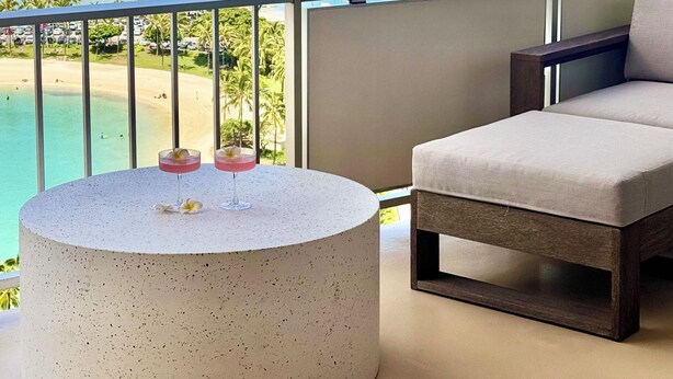 A balcony with a round table and a cushioned chair, overlooking a beach and ocean.