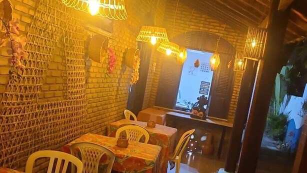 Un restaurant accueillant aux murs en briques, aux meubles en osier et à l’éclairage chaleureux. Les tables sont recouvertes de nappes à motifs floraux, et des lampes en osier sont suspendues au plafond.