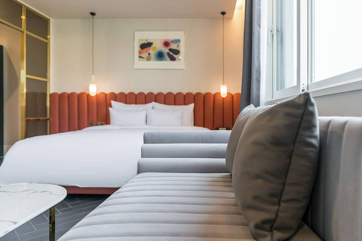 A modern hotel room with a large bed, a sofa, a small table, and a window with curtains.