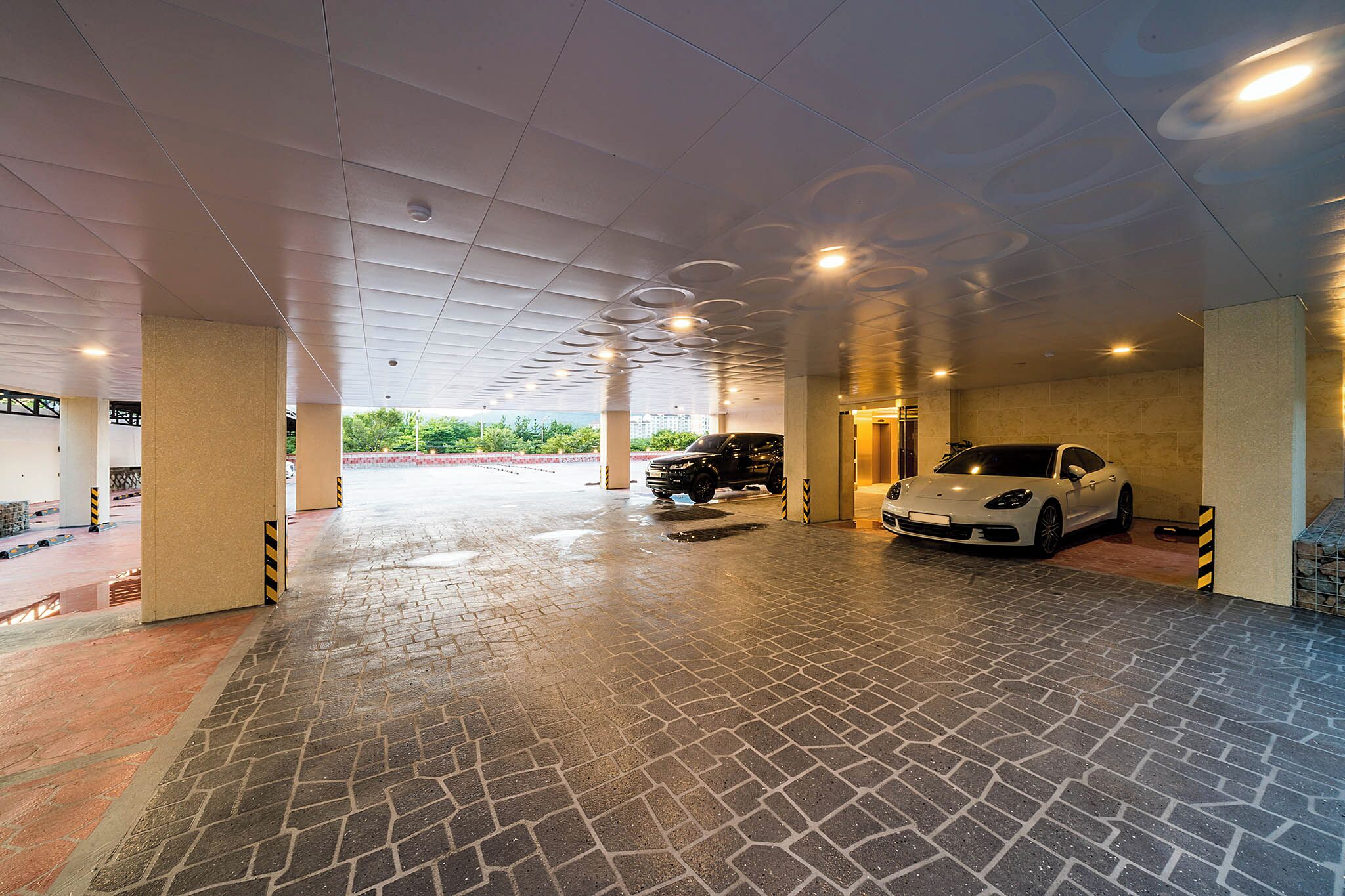 A covered parking area with a paved floor, two cars parked, and a view of greenery outside.