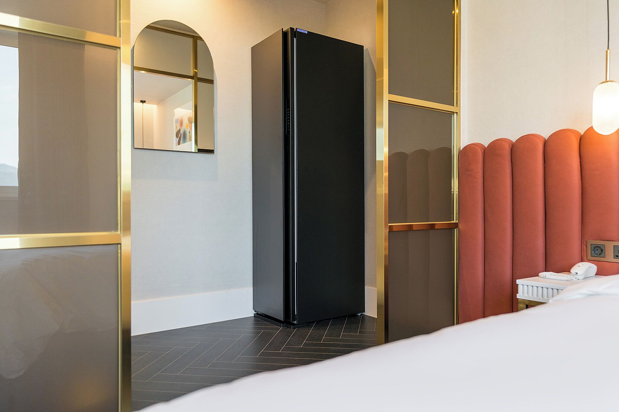 A modern hotel room with a black minibar refrigerator, a bed with white bedding, a bedside table with a lamp, and a red accent wall.