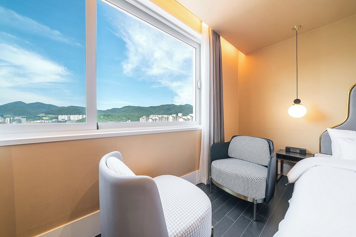 A hotel room with a large window, a bed, a chair, a bedside table, and a lamp.