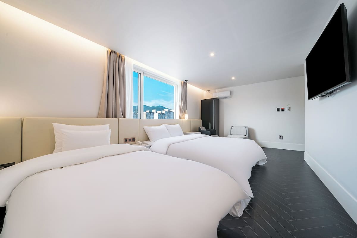 A hotel room with two beds, a large window with curtains, a flat-screen TV mounted on the wall, and an air conditioning unit.