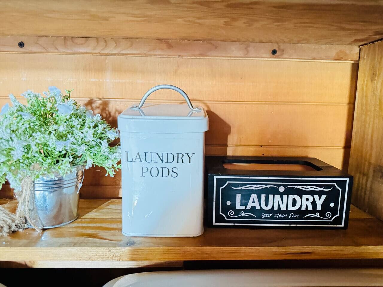 Ein Lagerraum für Wäsche mit einem Metallbehälter, beschriftet mit „LAUNDRY PODS“, und einer schwarzen Kiste mit derselben Beschriftung.