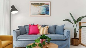 Ein Wohnzimmer mit einem blauen Sofa, einem runden Holztisch, einer Stehlampe und einem Bild von einer Strandlandschaft.
