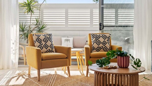 Ein modernes Wohnzimmer mit zwei senffarbenen Sesseln, einem runden Couchtisch und einem kleinen Holztisch. Die Sessel sind mit gemusterten Kissen ausgestattet und auf dem Couchtisch steht ein Blumentopf.