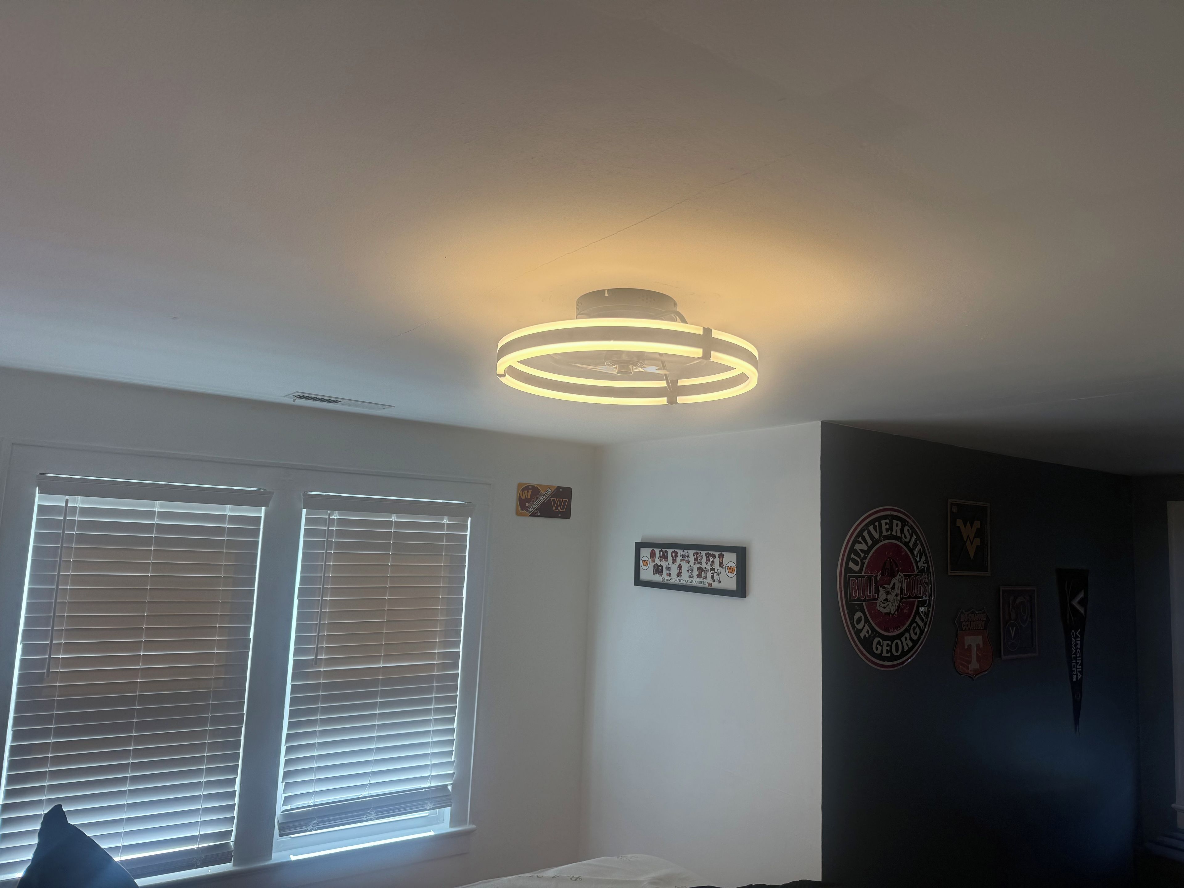 A room with a ceiling light fixture, a window with blinds, and a wall with various framed items.