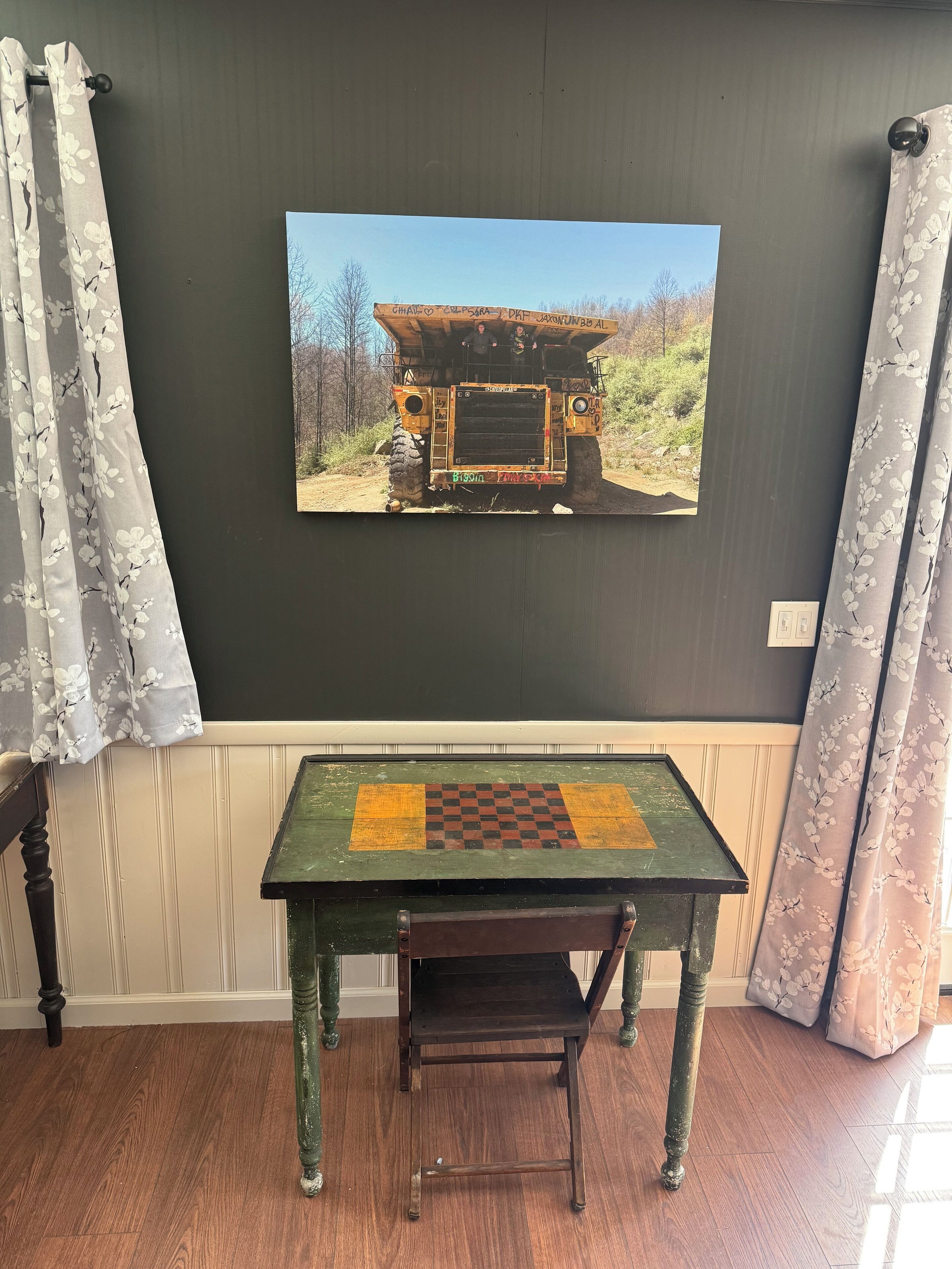 Una habitación con una pared oscura, un cuadro enmarcado de un vehículo, una mesa verde con un tablero de ajedrez y cortinas con estampado floral.