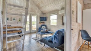 L’intérieur chaleureux d’un chalet avec un lit en mezzanine, un fauteuil bleu, une table à manger en bois et un téléviseur fixé au mur.