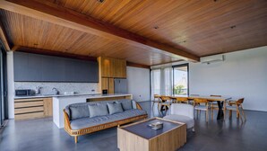 Una cocina moderna con armarios de madera y encimera gris, un área de comedor con mesa y sillas de madera, y un salón con sofá gris y mesa de centro de madera.