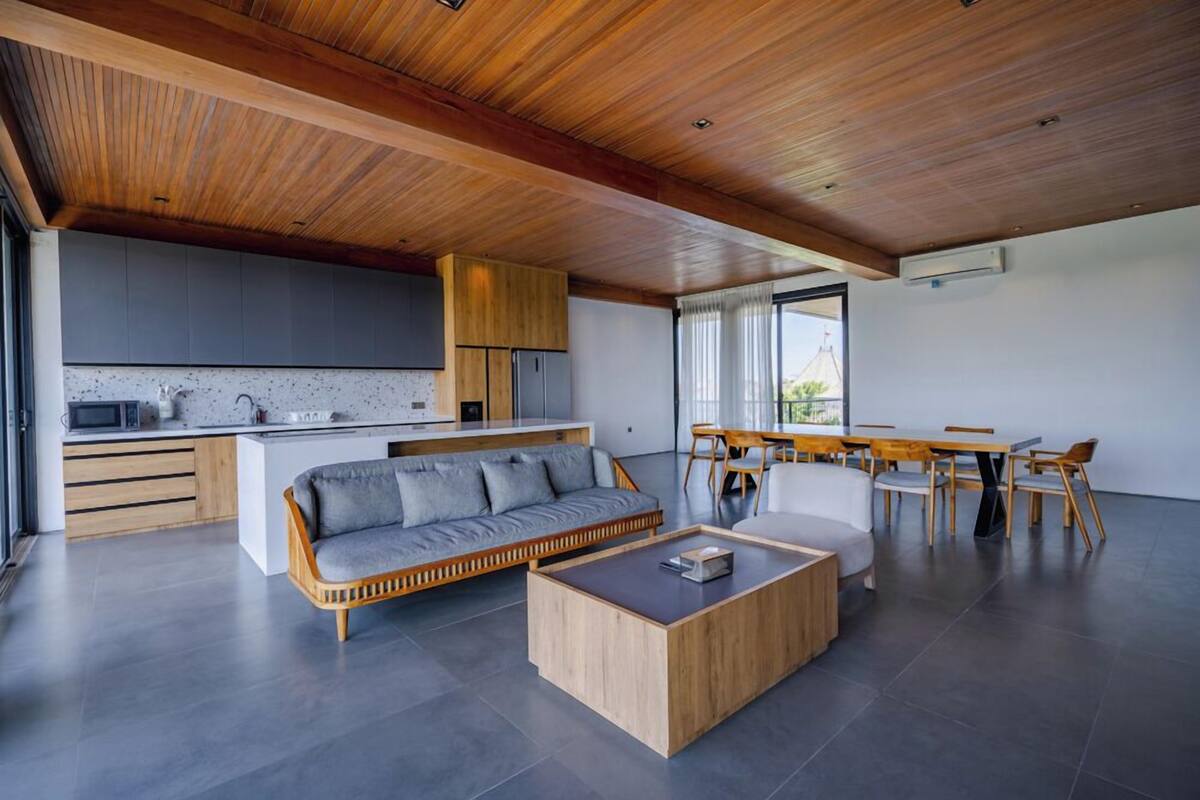 Una cocina moderna con armarios de madera y encimera gris, un área de comedor con mesa y sillas de madera, y un salón con sofá gris y mesa de centro de madera.