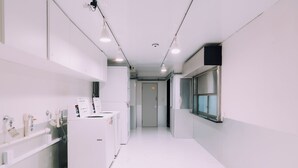 A clean, modern hallway with white walls and floors, featuring a laundry area with industrial-style machines, a window on the right, and a door at the end. - Minimalist Loft 1002 (Osaka)