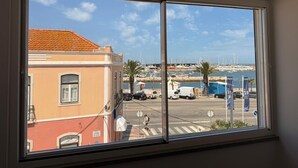 Vista desde una ventana a una zona costera con un puerto deportivo, vehículos estacionados y un edificio con techo rojo.