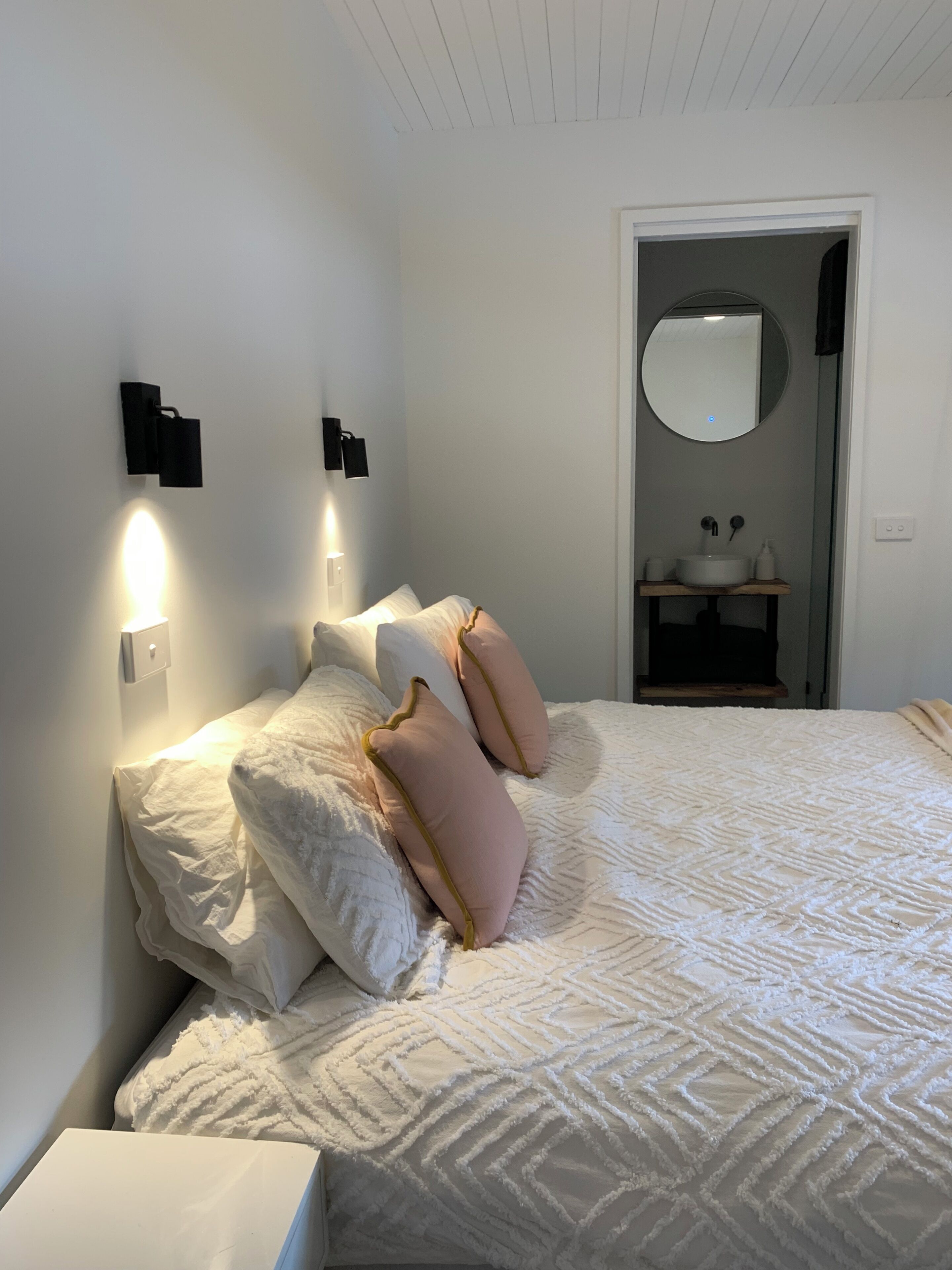 A bedroom with a bed, pillows, a nightstand, a wall-mounted light, a mirror, and a door leading to a bathroom.