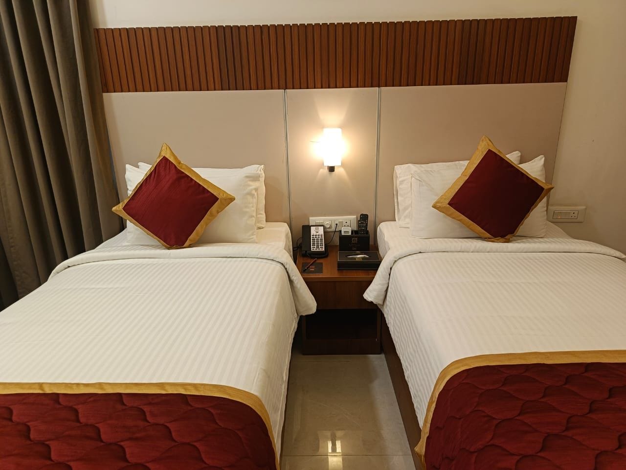 A hotel room with two beds, a nightstand with a phone and a lamp, and a window with curtains.