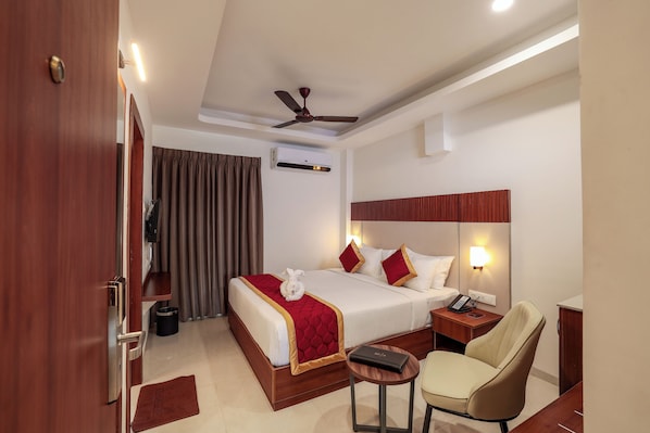 A hotel room with a bed, a chair, a bedside table, a lamp, and a ceiling fan. - Sathyam Oasis (Tiruchirappalli)