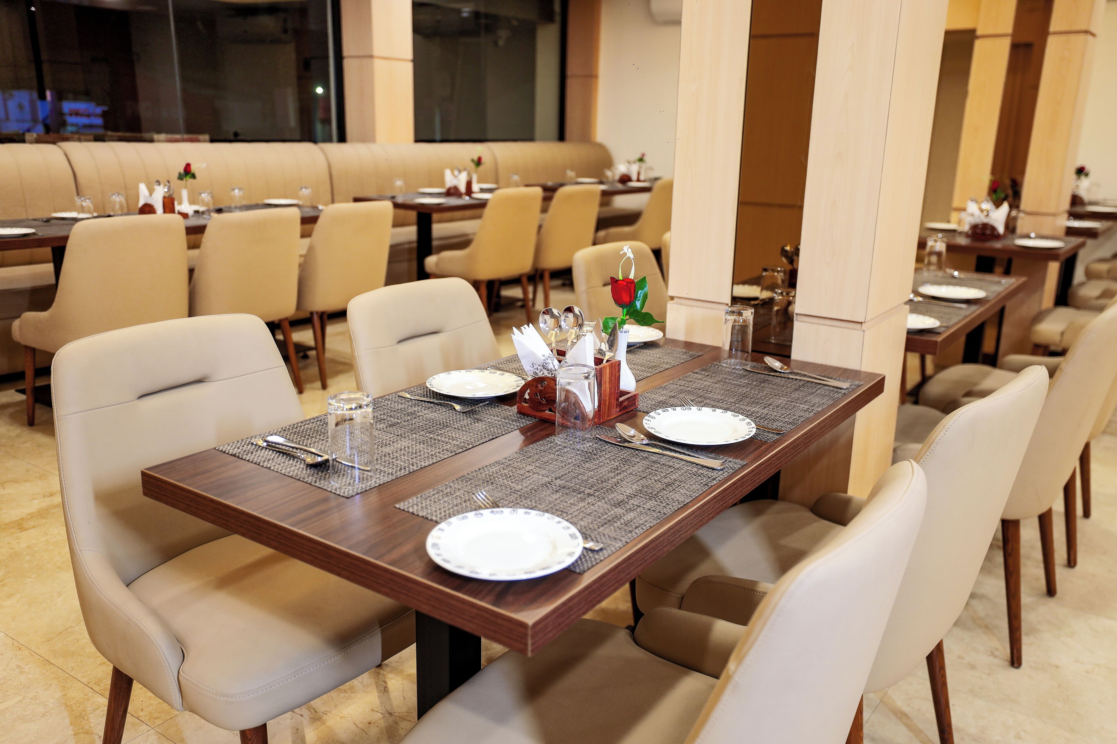 A dining area with tables set for four, each with a placemat, plates, cutlery, and a vase with a single red rose.