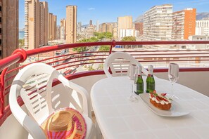 A balcony with white chairs, a table with a dessert and wine bottles, and a cityscape view.