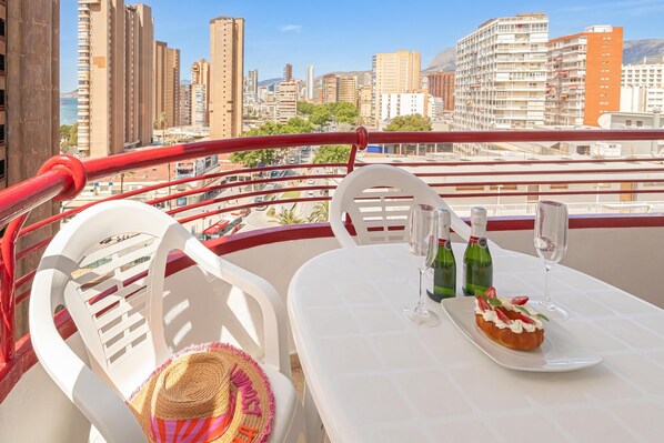 A balcony with white chairs, a table with a dessert and wine bottles, and a cityscape view.