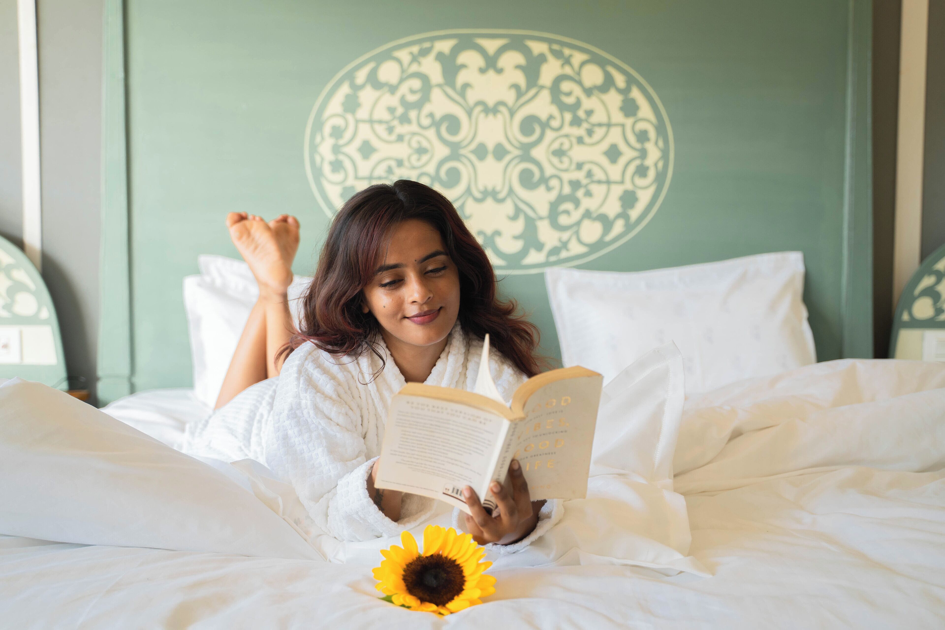 A person lying on a bed with a book and a sunflower, in a room with a decorative wall pattern.