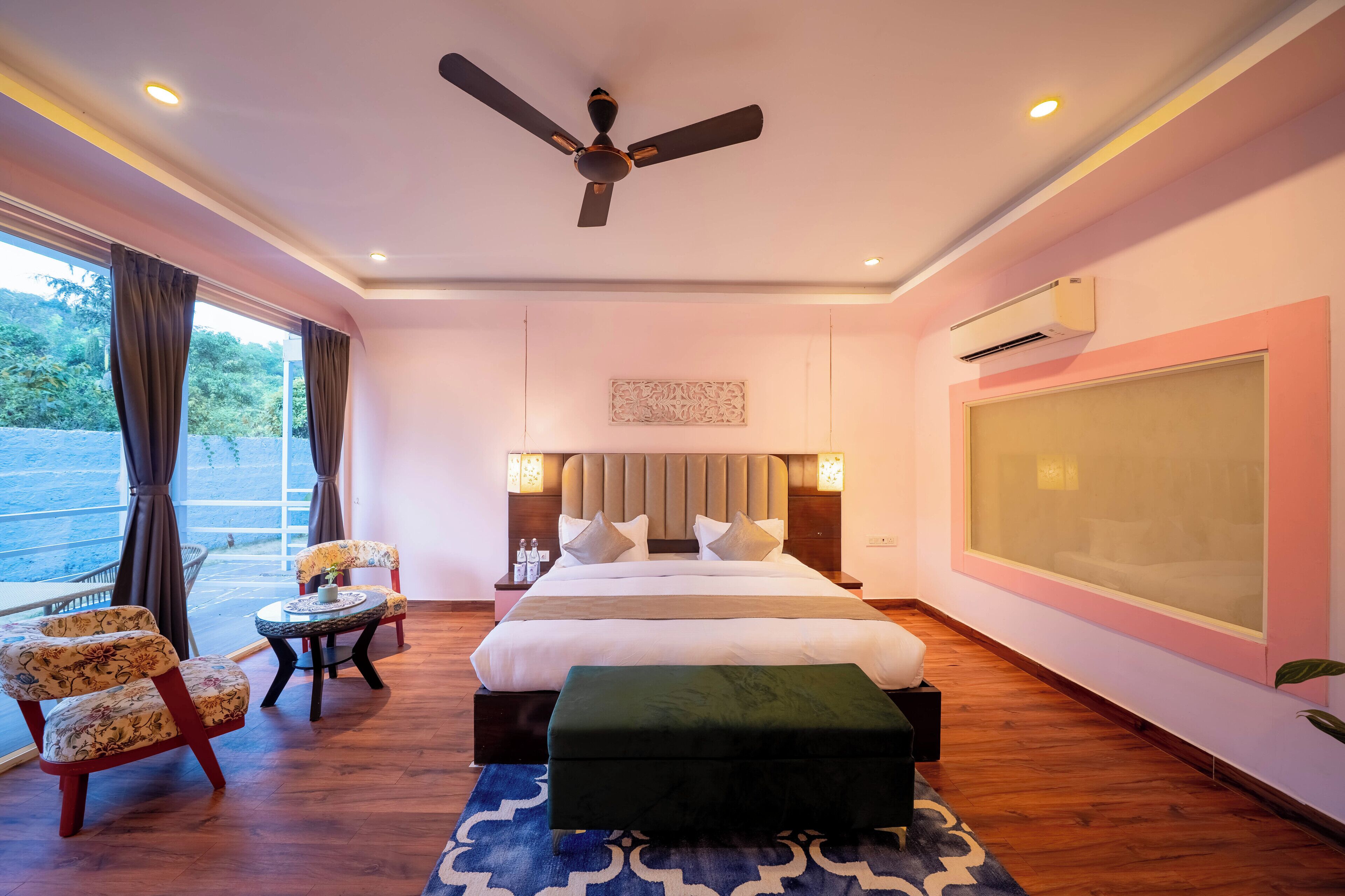 A hotel room with a large bed, a ceiling fan, a large window with curtains, a small round table, a chair, and a large mirror.