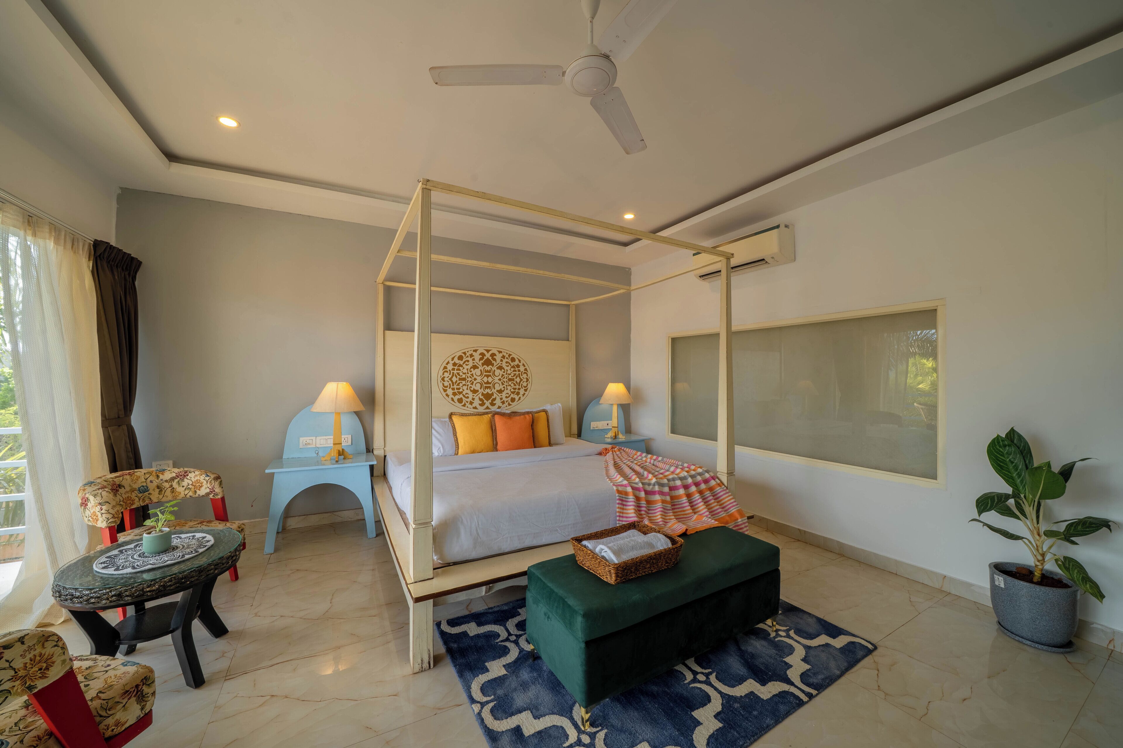 A four-poster bed with a canopy, a nightstand with lamps, a chair with a plant, a small table with a decorative item, and a potted plant.