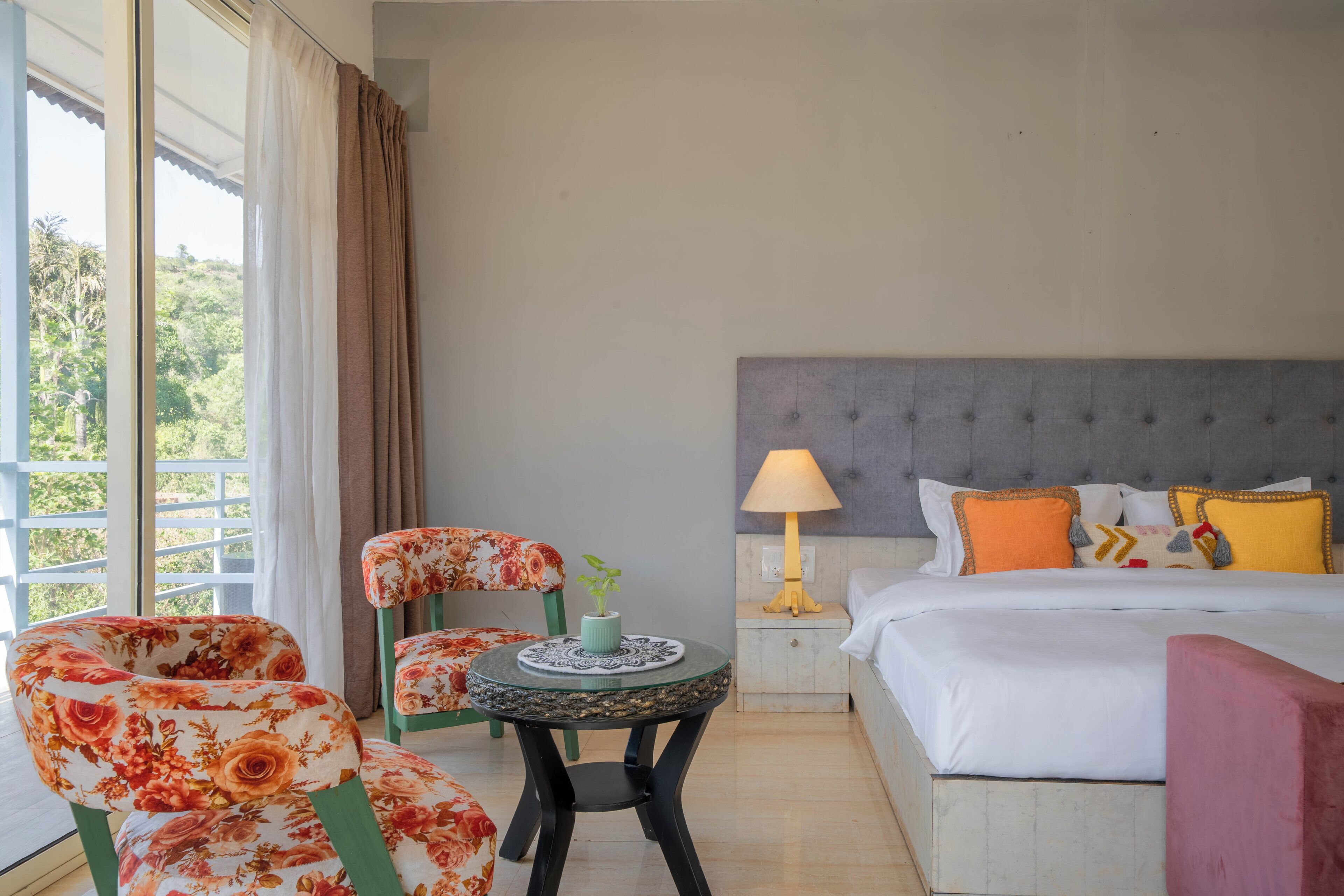 A hotel room with a bed, a nightstand, a lamp, a chair, and a small table with a plant.