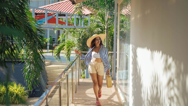 A person walking on a sunlit outdoor corridor with a railing, palm trees, and a building with a red roof in the background. - Perfectstayz Koko Maya (Morjim)