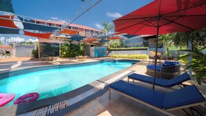 A swimming pool with lounge chairs and umbrellas, surrounded by a building with a red roof and a view of palm trees. - Perfectstayz Koko Maya (Morjim)