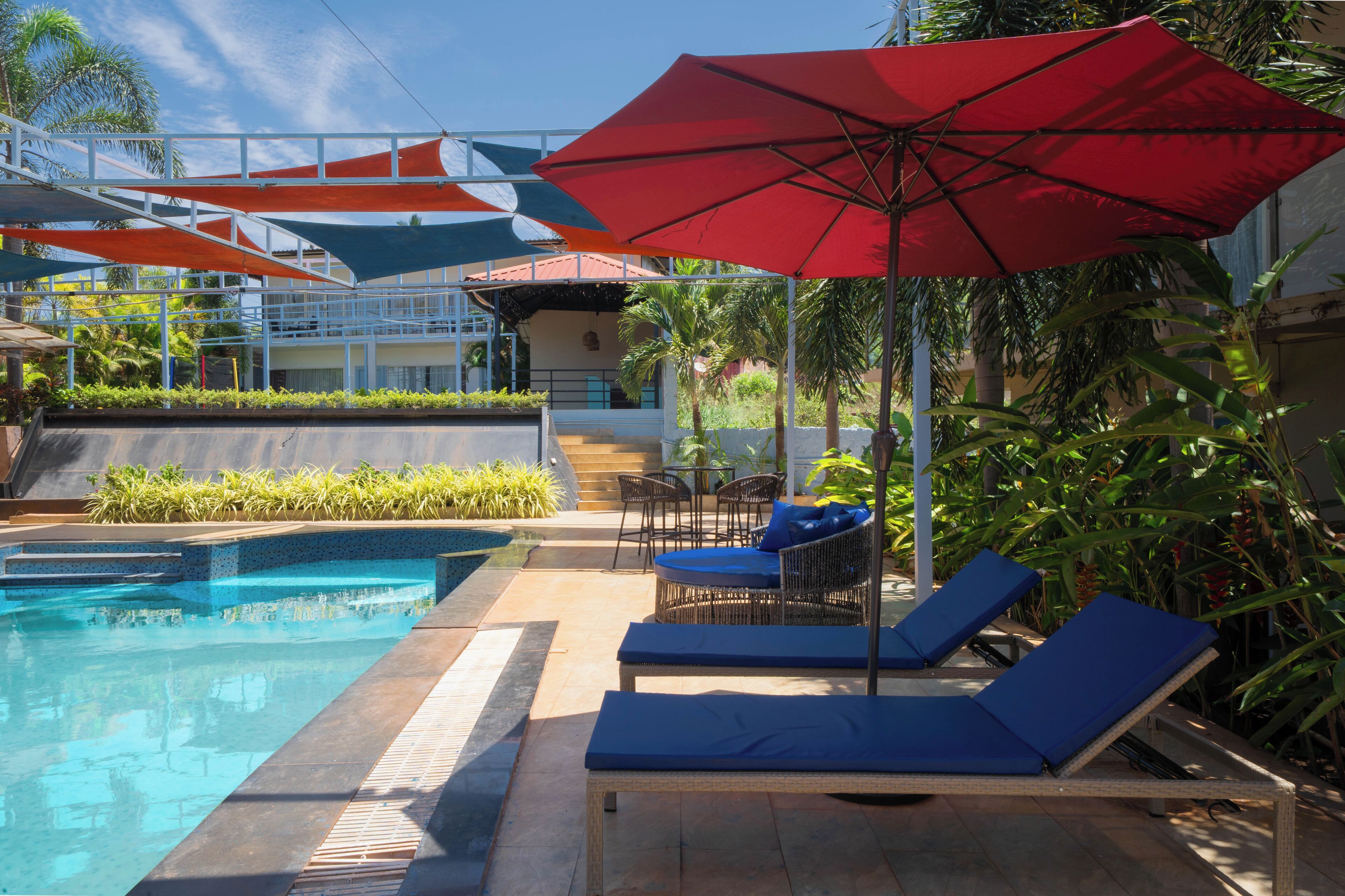 Un espace piscine avec des chaises longues et des parasols, entouré de plantes et avec un bâtiment en arrière-plan.