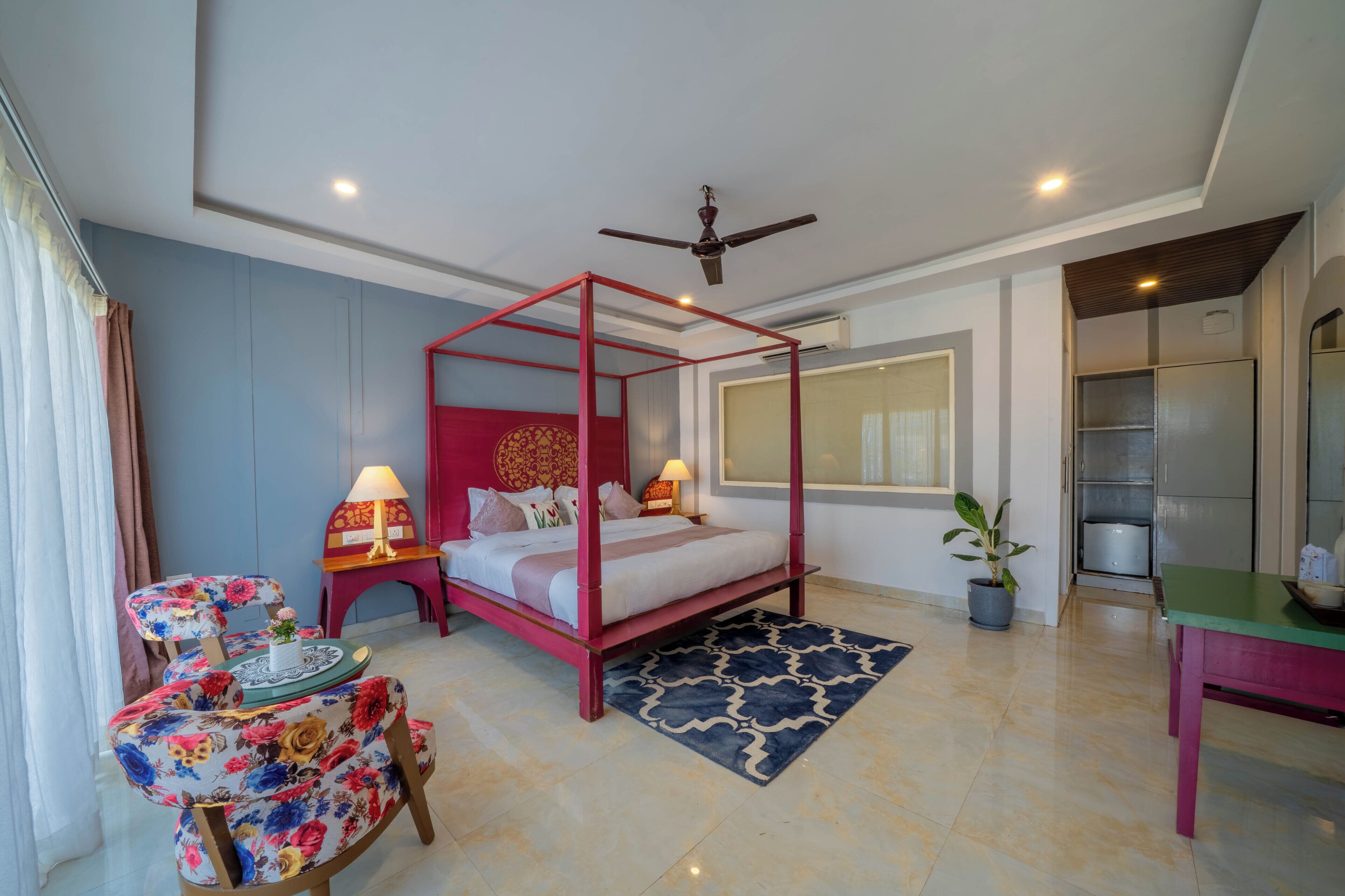 A four-poster bed with a ceiling fan, a floral armchair, a small table with a lamp, and a potted plant.