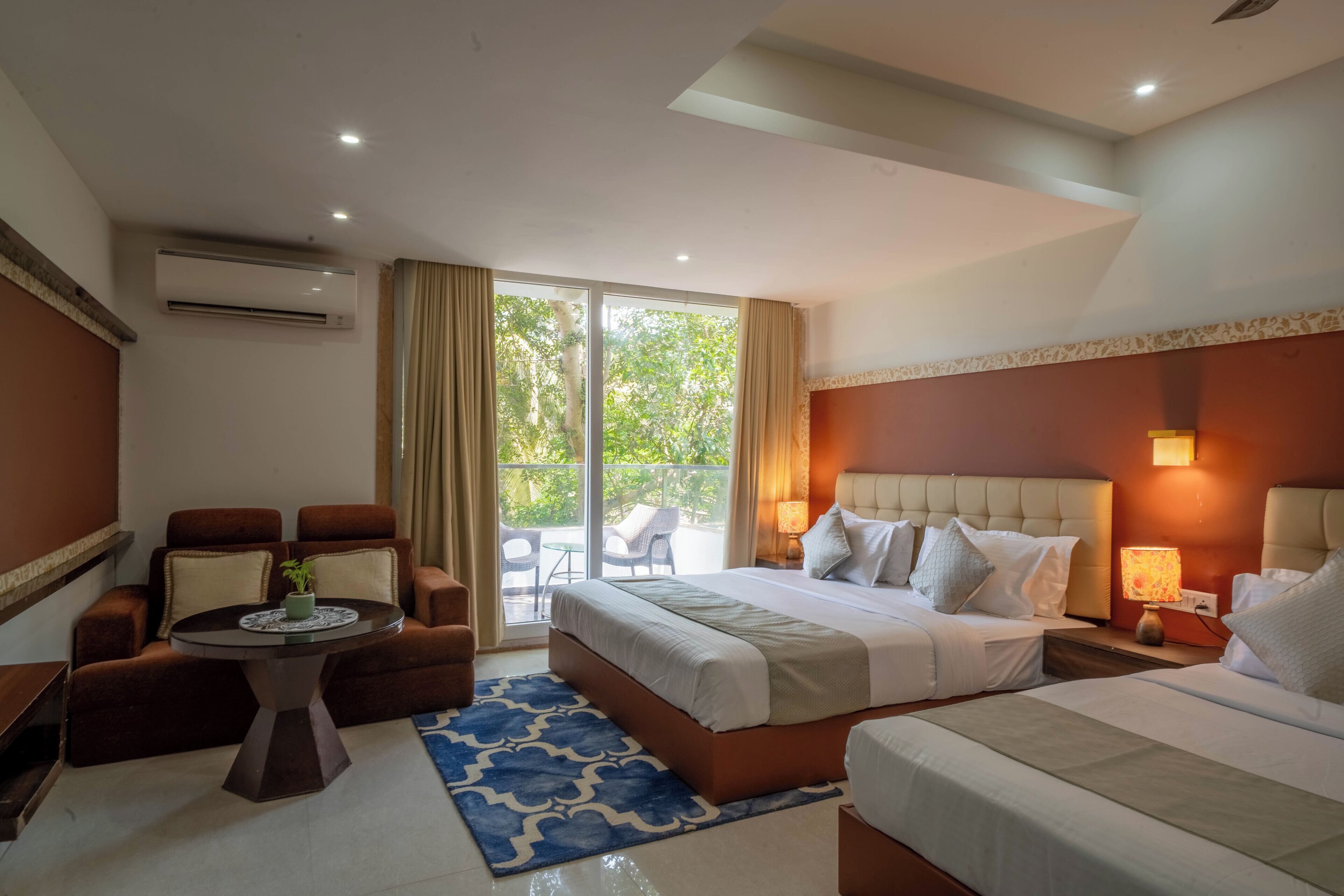 A hotel room with a large bed, a sofa, a small table, and a balcony with outdoor furniture.