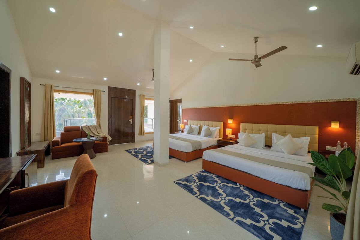 A hotel room with two beds, a sofa, a dining table, and a ceiling fan.