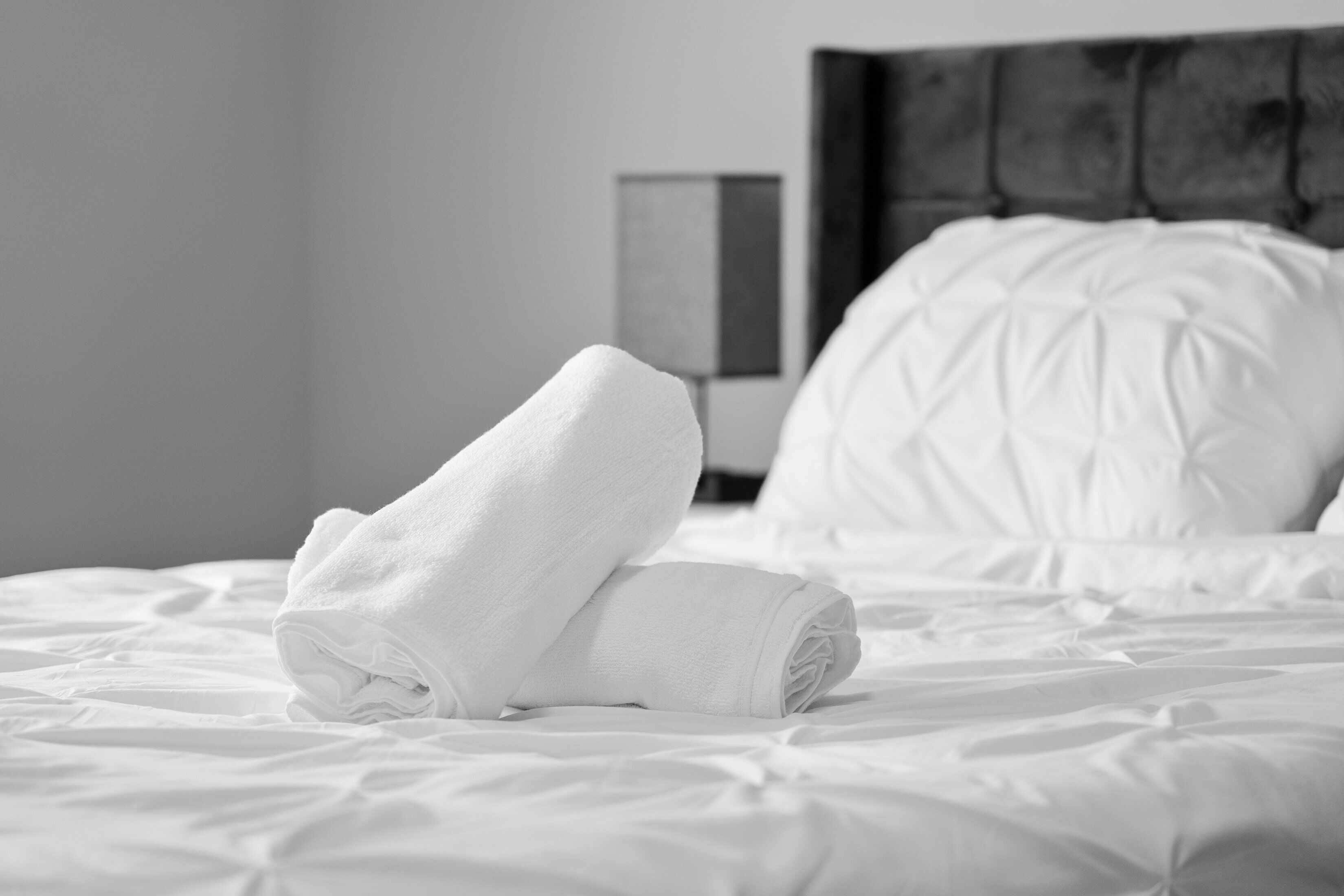 Una cama bien hecha con sábanas blancas y una toalla enrollada encima, un cabecero y una mesita de noche con una lámpara.