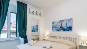 A hotel room with a bed, a window with curtains, an air conditioner, a painting of sailboats, and a bedside table with a lamp. - Galatea by Interhome (Sanremo)