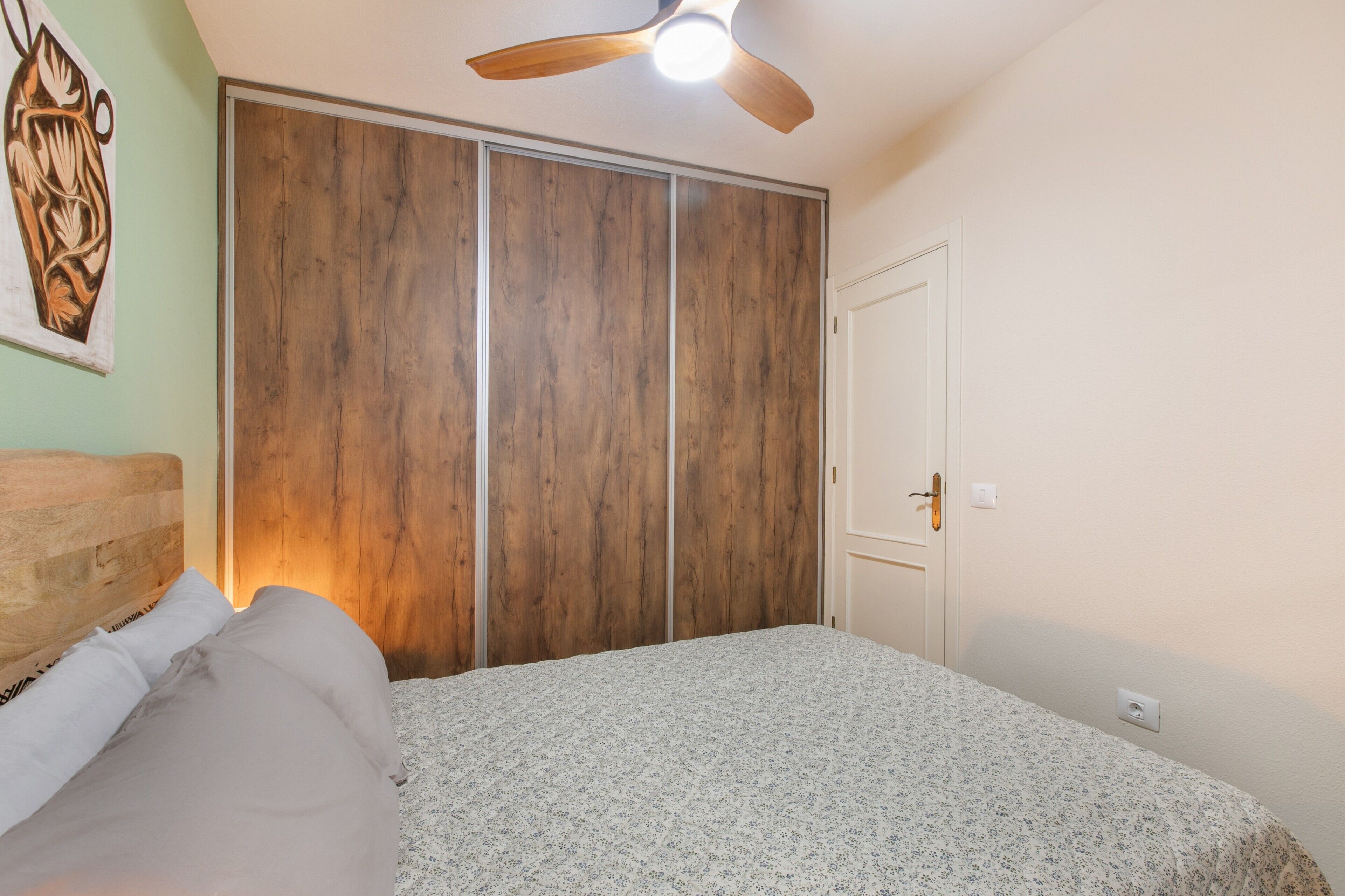 A bedroom with a bed, wooden wardrobe, ceiling fan, and a wall-mounted artwork.