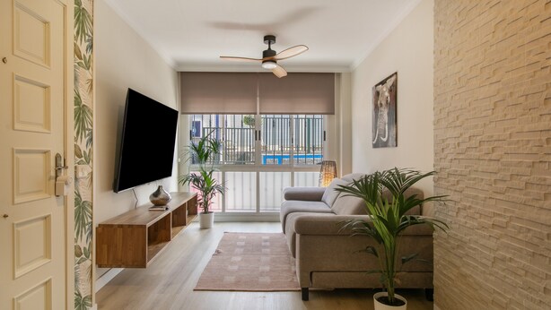 Een woonkamer met een flatscreen televisie, een plafondventilator, een bank, een kamerplant en uitzicht op een zwembad door een raam.
