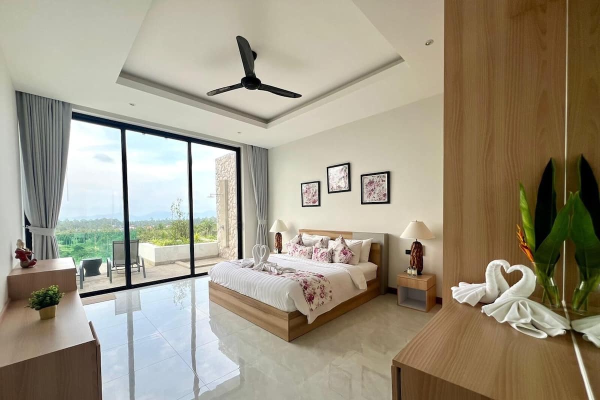 A modern bedroom with a large bed, a ceiling fan, a view of greenery through large windows, and a wooden nightstand with a lamp.