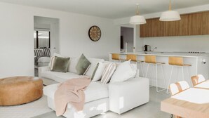 Una sala de estar moderna con un sofá blanco, un reposapiés rosa y una zona de cocina con armarios de madera y taburetes de bar.