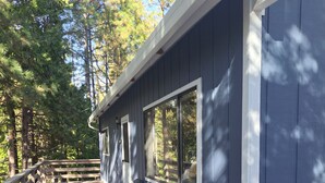 A covered wooden deck with a dark blue exterior wall, large windows, and a wooden railing. -    Cozy cottage for vacation in Cedar Ridge near its famous trail. (Sonora)