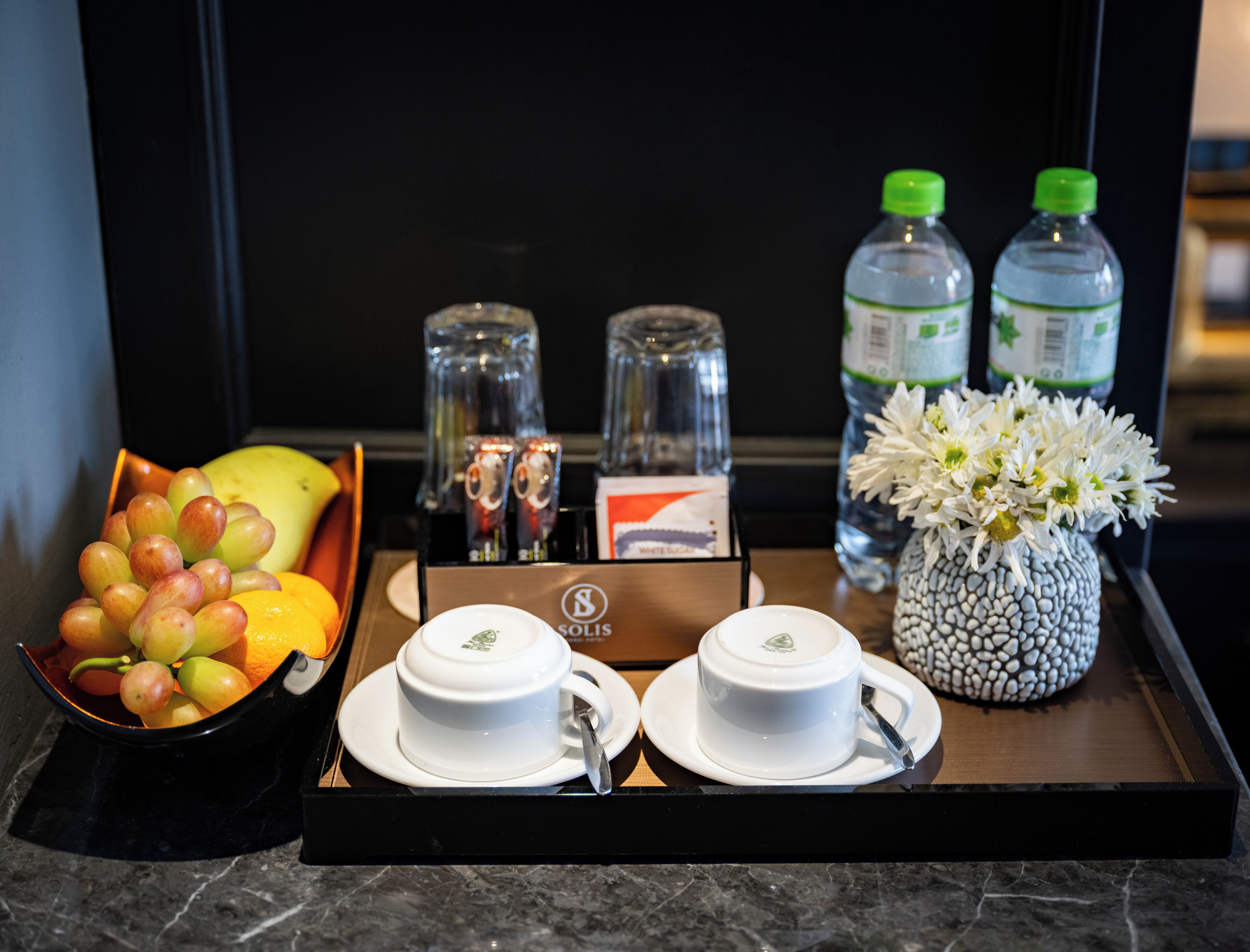 A tray with a fruit bowl, two cups, a vase with flowers, and two water bottles.