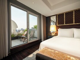 A hotel room with a large bed, a balcony with chairs and plants, and a view of the cityscape.